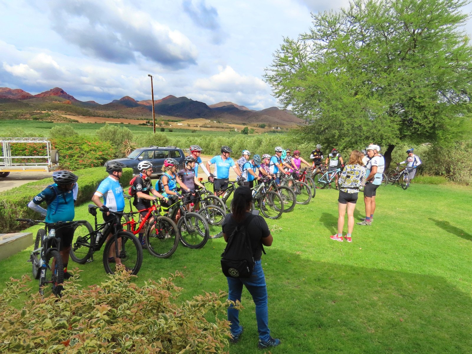 Group of people wearing biking gear and helmets standing with their bikes on a grassy area, listening to a woman who appears to be giving instructions, with a large green tree, a car, mountains, and a cloudy sky in the background.