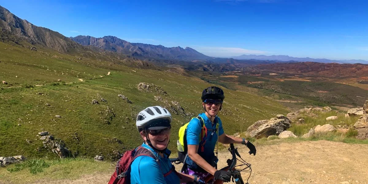 Two women in cycling gear and helmets smiling while riding mountain bikes on a trail in a mountainous landscape with green hills and a clear blue sky.