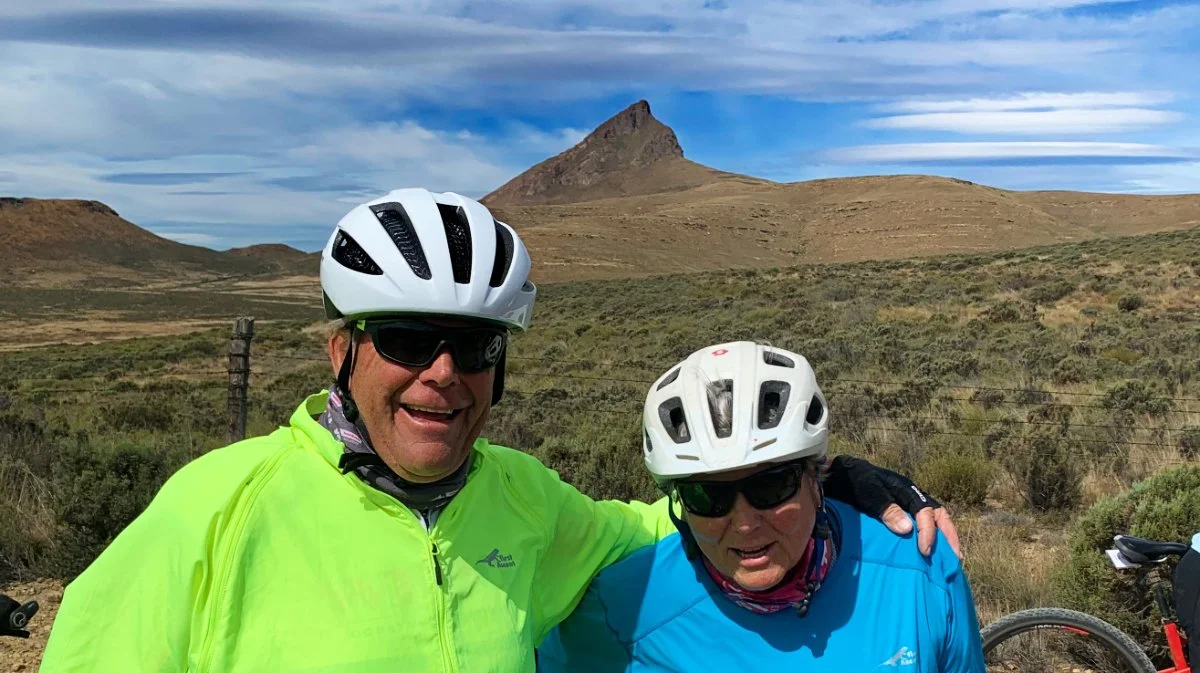Two cyclists wearing helmets and sunglasses outdoors, with a mountainous landscape in the background.