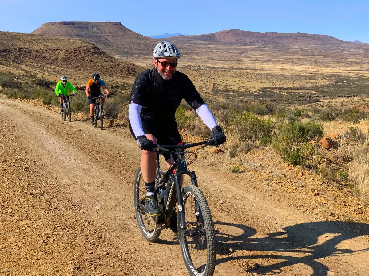 A group of four mountain bikers riding on a dirt trail through a desert landscape under a clear blue sky, with mountains in the background.