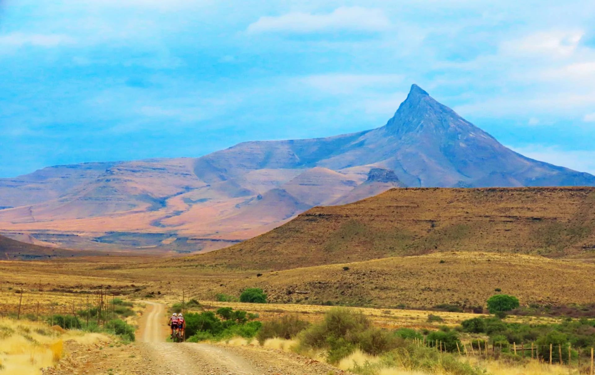 Hikers walking along a dirt trail in a vast, arid landscape with mountains, including a sharply pointed peak, in the background under a partly cloudy sky.