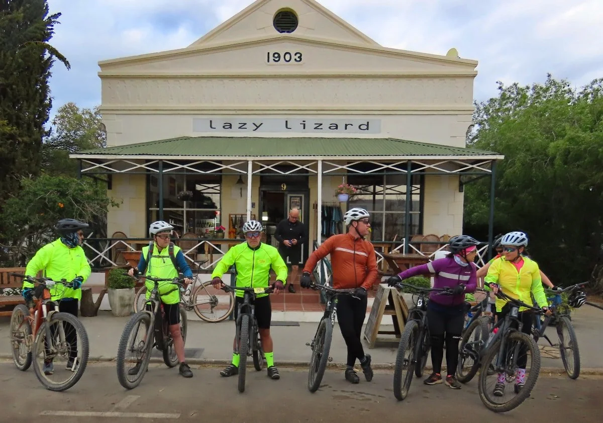 Group of six cyclists in high-visibility clothing and helmets standing with their bicycles in front of a café named Lazy Lizard, built in a historic style with a white facade and green roofing, with a person sitting inside and trees surrounding the a