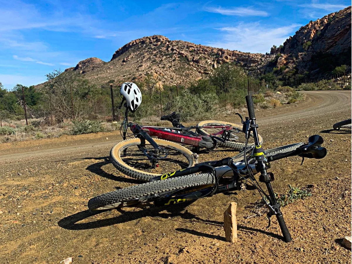 Two mountain bikes lying on a dirt trail in a desert landscape with hills and sparse vegetation, and a blue sky.