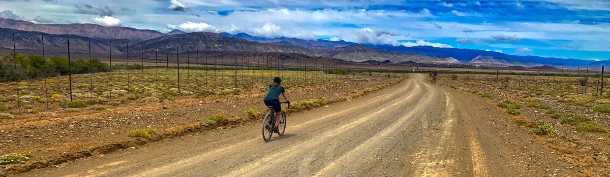 A person riding a bike on a dirt road through an arid landscape with mountains in the distance under a partly cloudy sky.