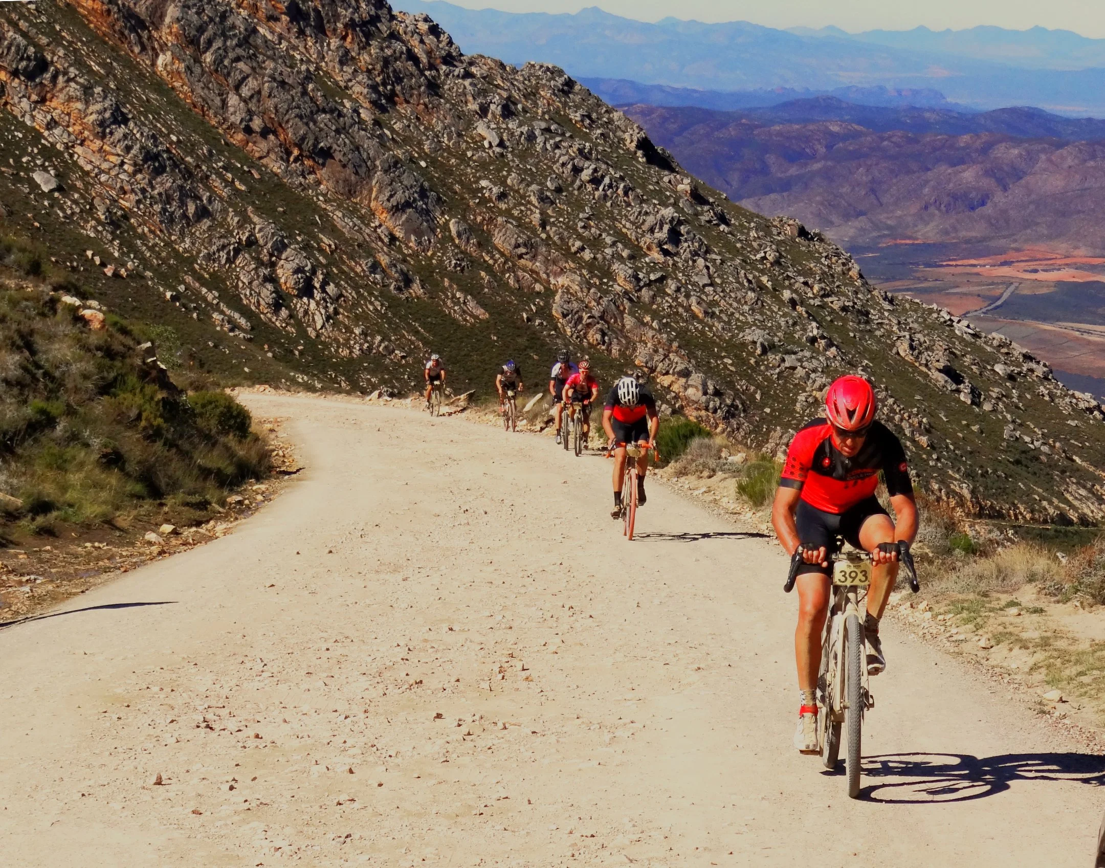 A group of cyclists riding on a dirt trail in a mountainous area, with rocky slopes and distant valleys visible in the background.