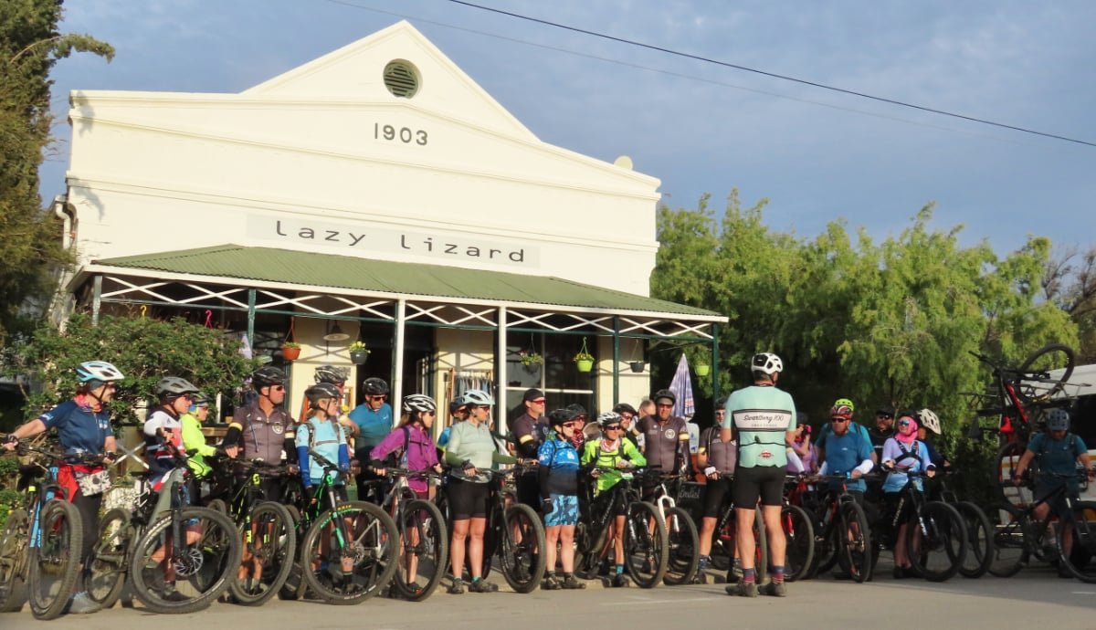Group of cyclists gathered in front of a building with a sign that reads "Lazy Lizard" and an older date, 1903.