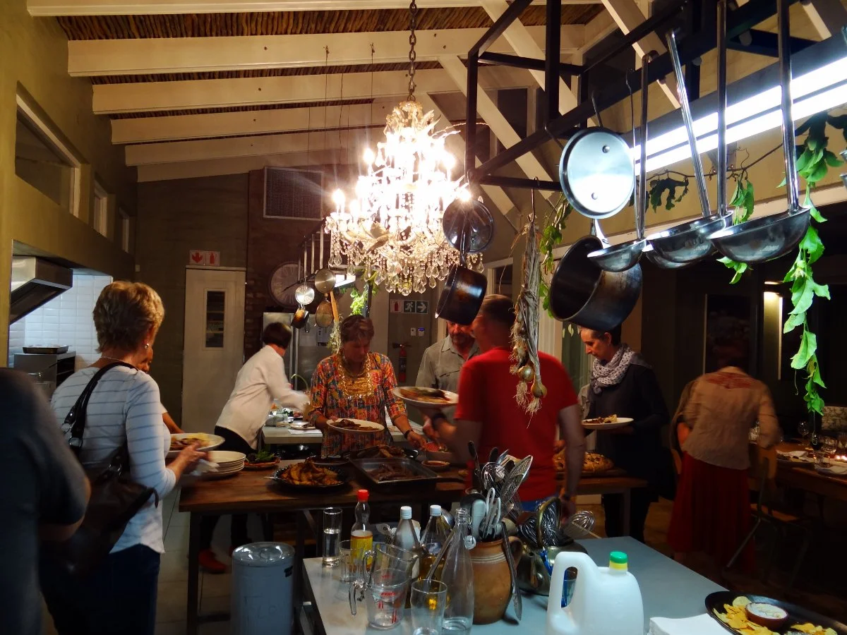 People serving food at a dinner party in a kitchen with hanging pots and pans, a chandelier, and guests standing around the table.