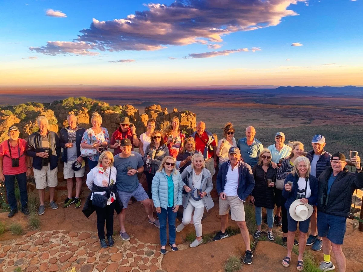 A group of around 20 people posing outdoors at sunset on a rocky overlook with a vast desert landscape in the background.