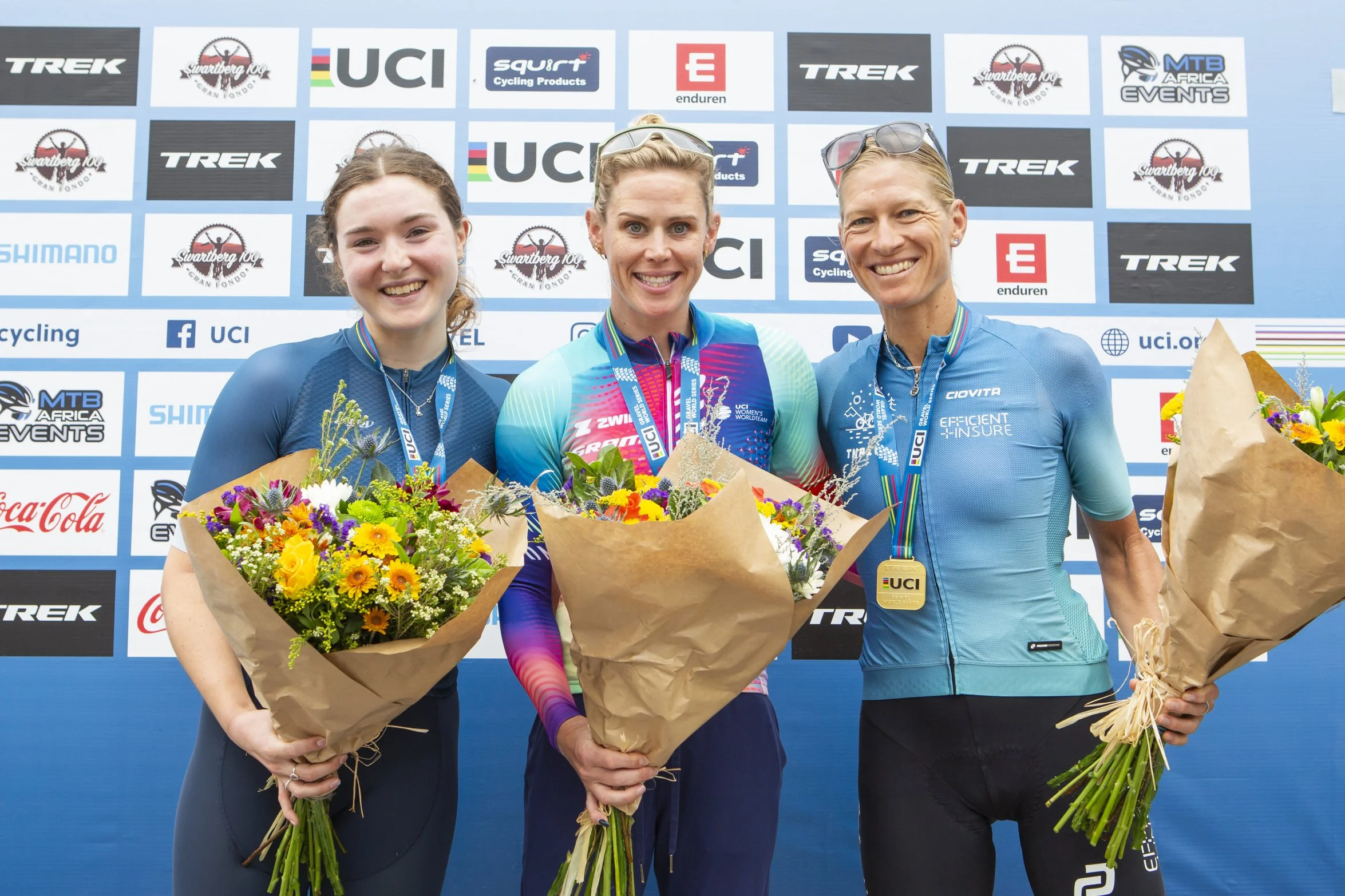 Three female cyclists on a podium holding bouquets of flowers, wearing medals and sporting cycling gear, with a backdrop displaying various sponsor logos and the UCI logo.