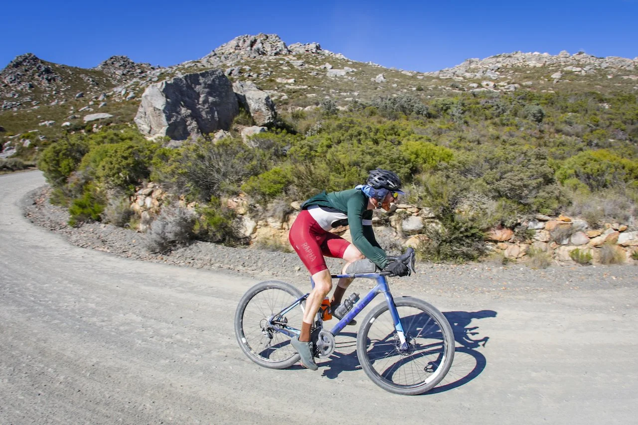 A cyclist riding a mountain bike on a dirt trail in a mountainous landscape with rocky hills and green shrubs under a clear blue sky.