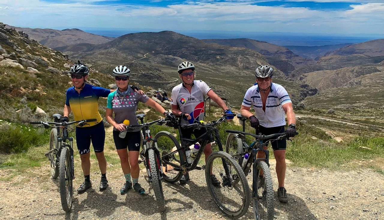 Four cyclists standing with bikes on a mountain trail with a scenic mountainous landscape in the background.