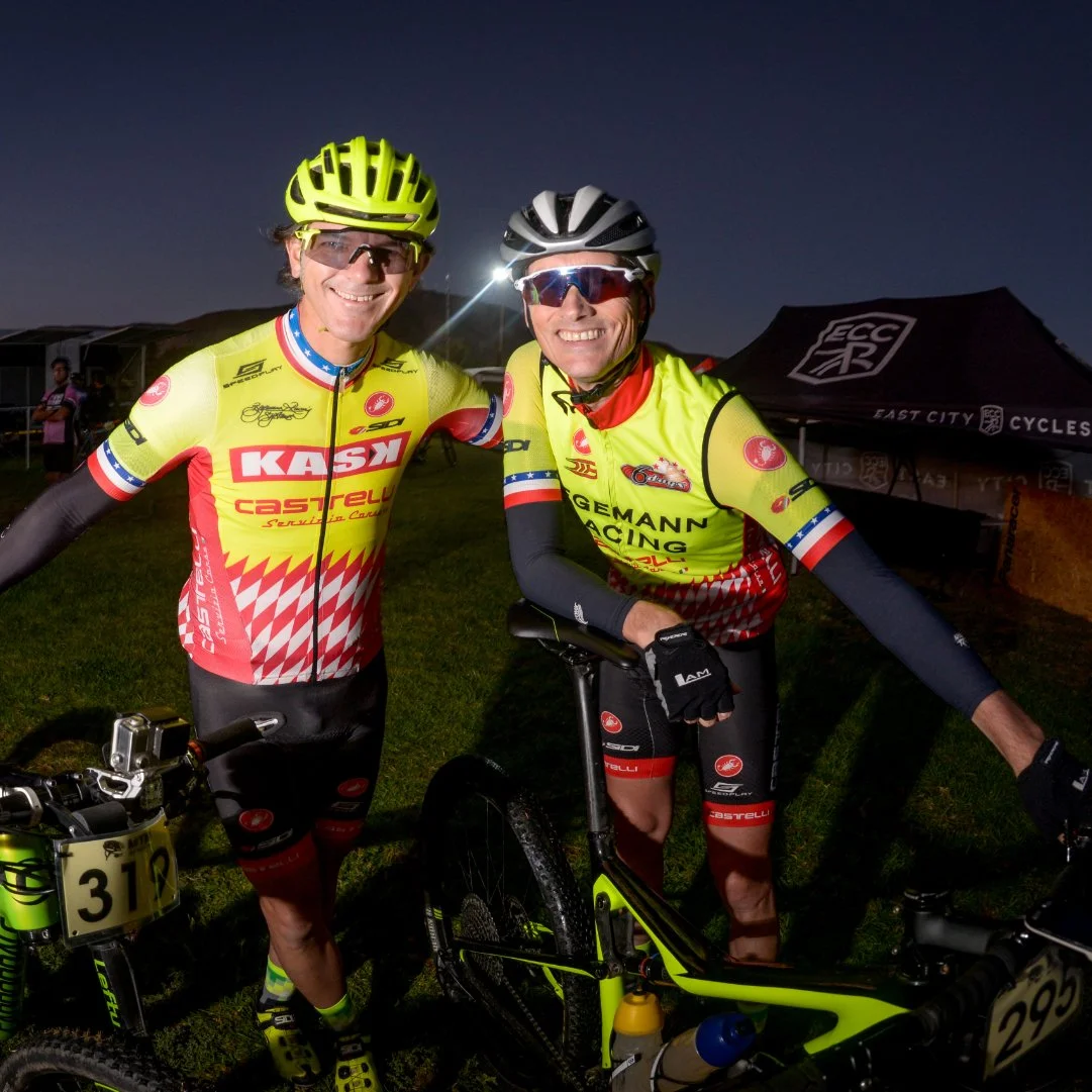 Two male cyclists dressed in yellow and red racing gear, wearing helmets and sunglasses, smiling and posing with their bikes at night.