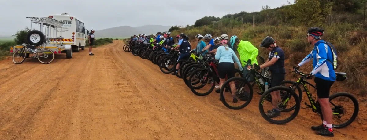 A group of mountain bikers standing with their bikes on a dirt road, preparing for a ride. There is a support vehicle and a bicycle on a trailer nearby. The background features hilly terrain and cloudy sky.