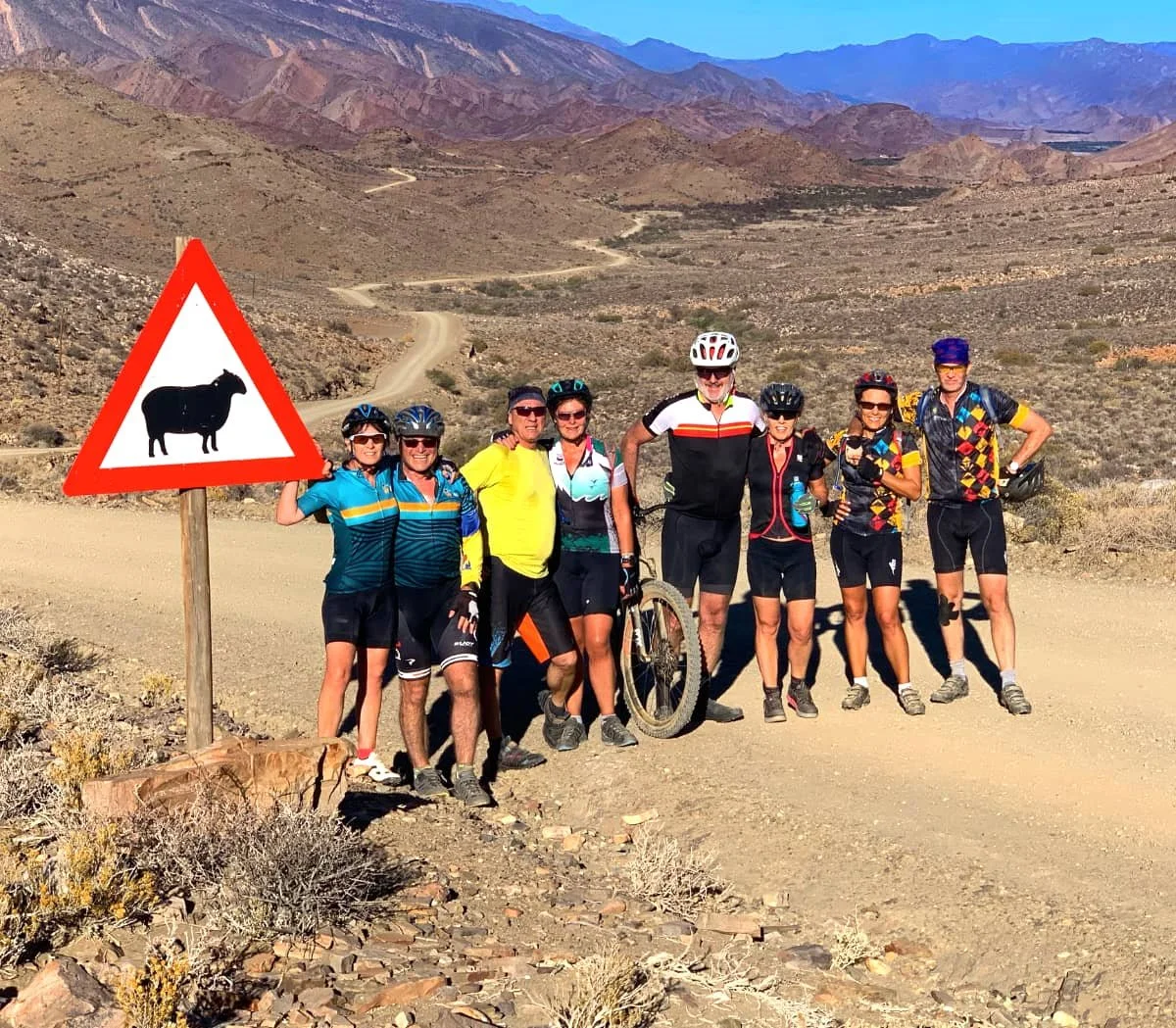 Group of eight cyclists standing on a dirt road in a desert landscape with mountains in the background. There is a cow crossing sign in the foreground.