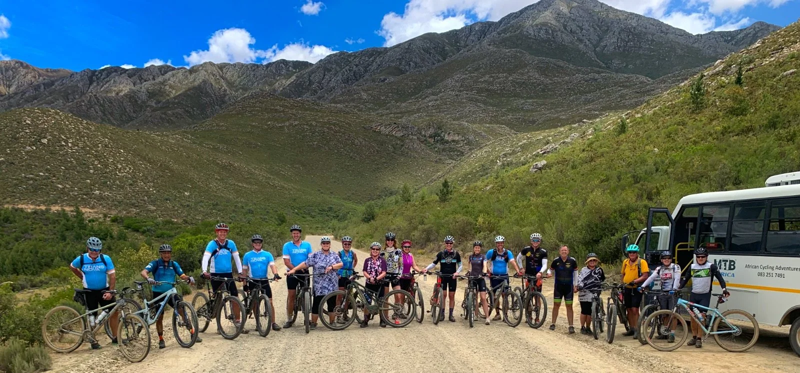 Group of 15 cyclists with their mountain bikes standing on a dirt trail in a mountainous landscape with green slopes and rocky peaks, accompanied by a white bus.