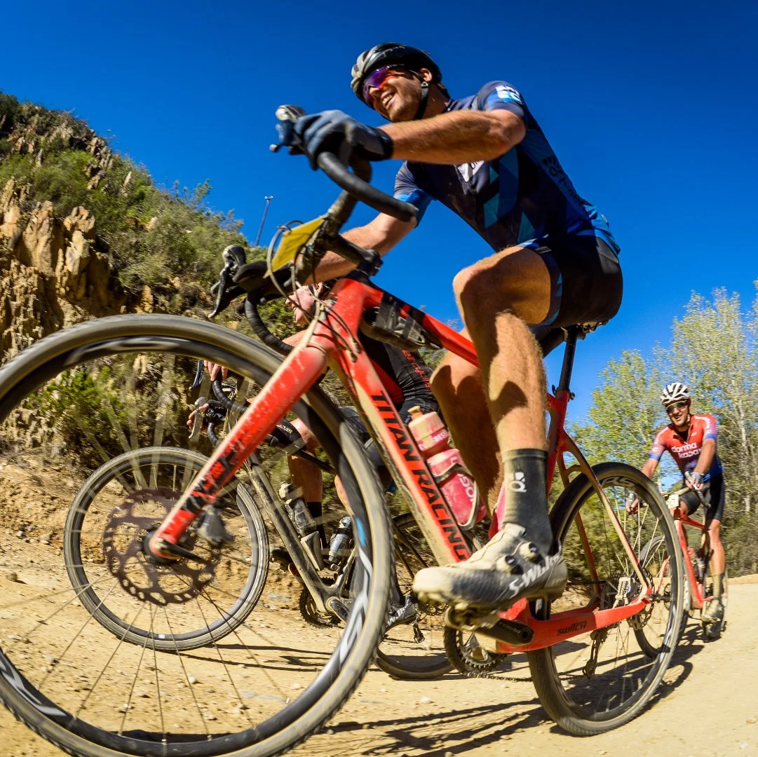 Group of cyclists riding mountain bikes on a dirt trail under a clear blue sky, with rocky hills and trees in the background.