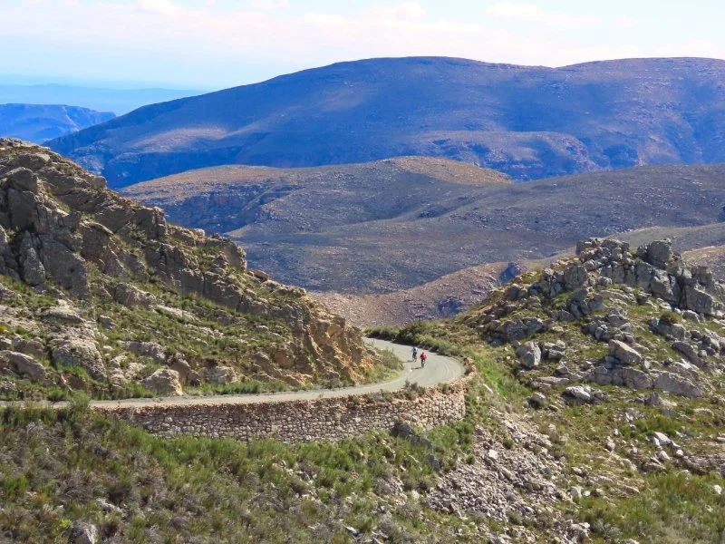 A winding mountain road with two cyclists riding on it, surrounded by rocky terrain and hills with large mountains in the background.
