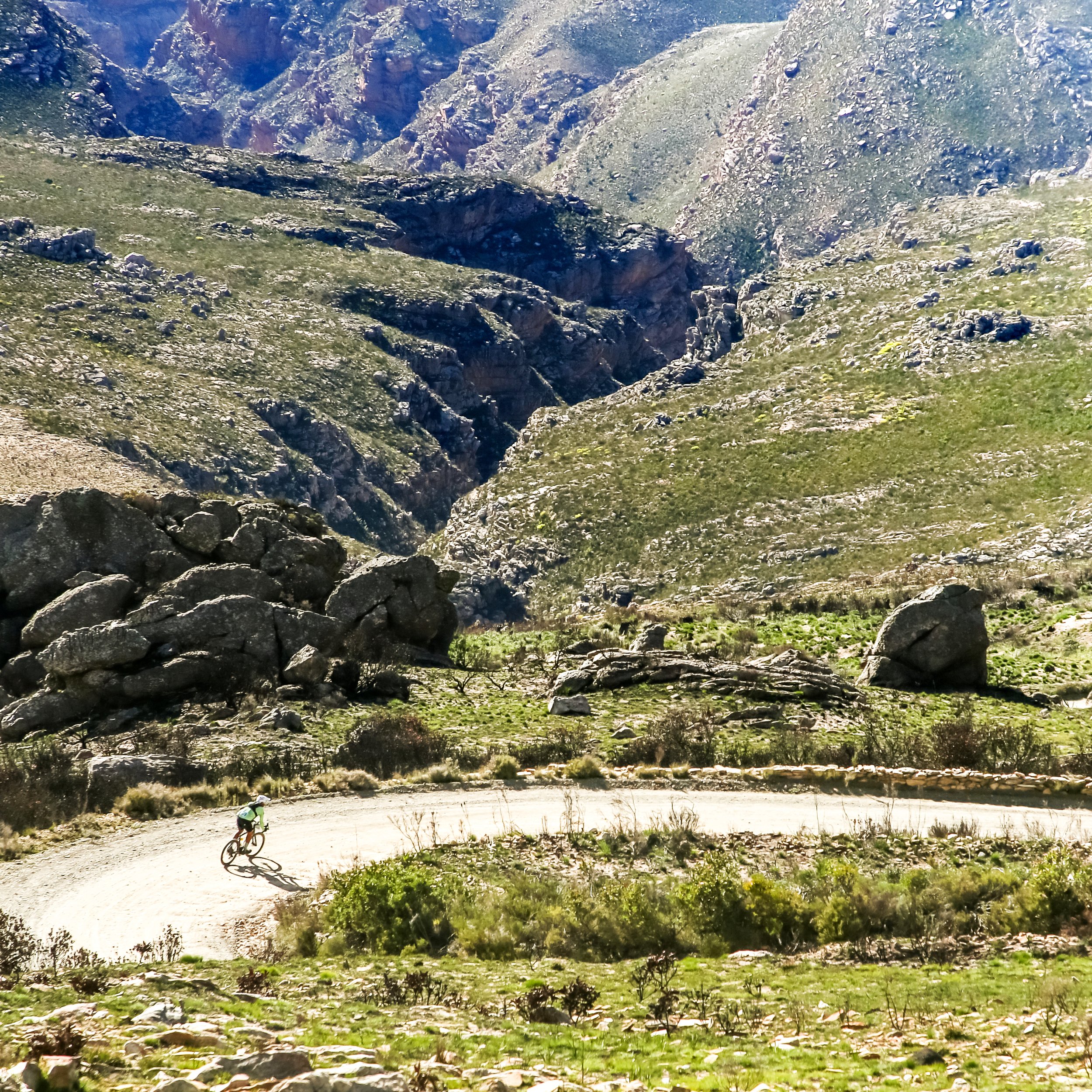 A cyclist riding on a winding dirt trail through a mountainous landscape with rocky slopes and sparse vegetation.