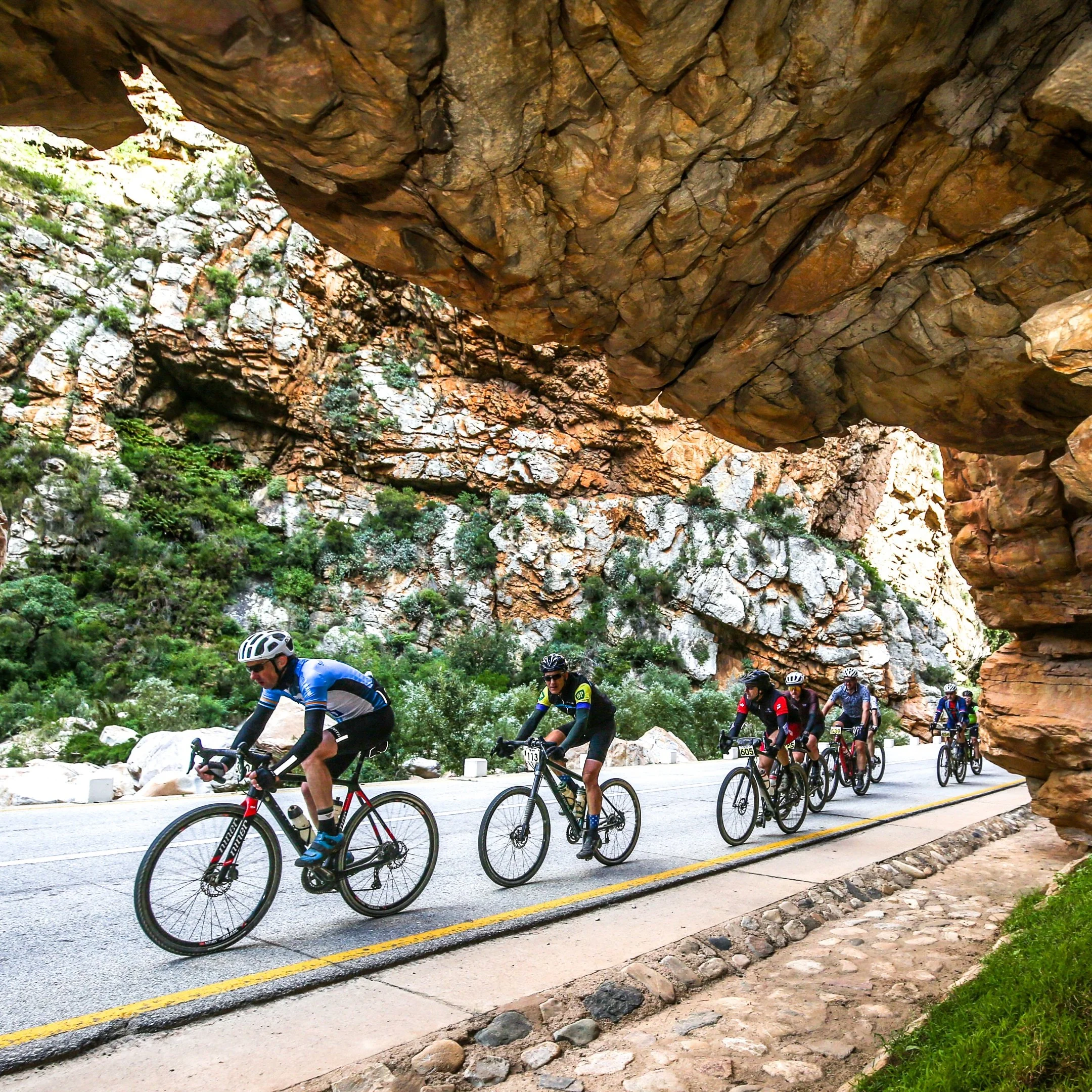 A group of cyclists riding on a mountain road passing beneath a large rocky overhang with lush green vegetation in the background.