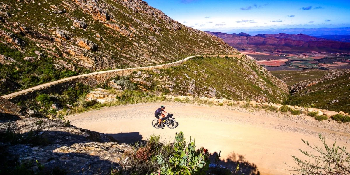 A person riding a mountain bike on a dirt trail in a mountainous landscape with steep hills and sparse vegetation under a partly cloudy sky.