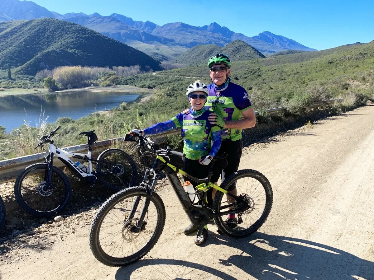 Two cyclists in colorful gear posing with bikes on a dirt trail in a mountainous landscape, with a lake and hills in the background on a clear, sunny day.