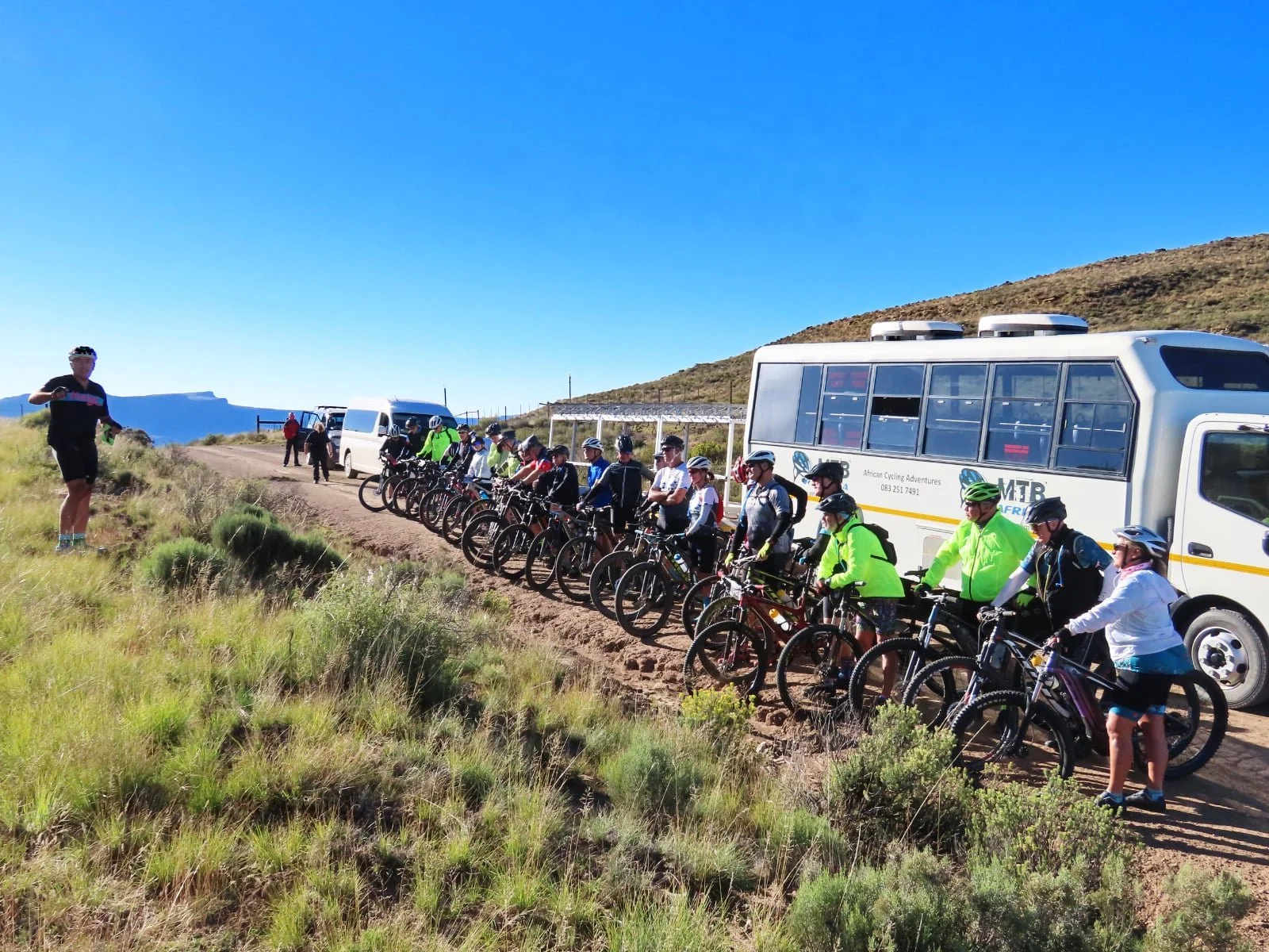 Group of cyclists with helmets and sports gear lined up beside a tour bus and vans on a dirt trail in a scenic, hilly landscape under clear blue sky.
