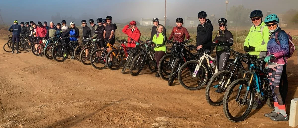 Group of people in cycling gear with bikes on a dirt trail in an open outdoor area.