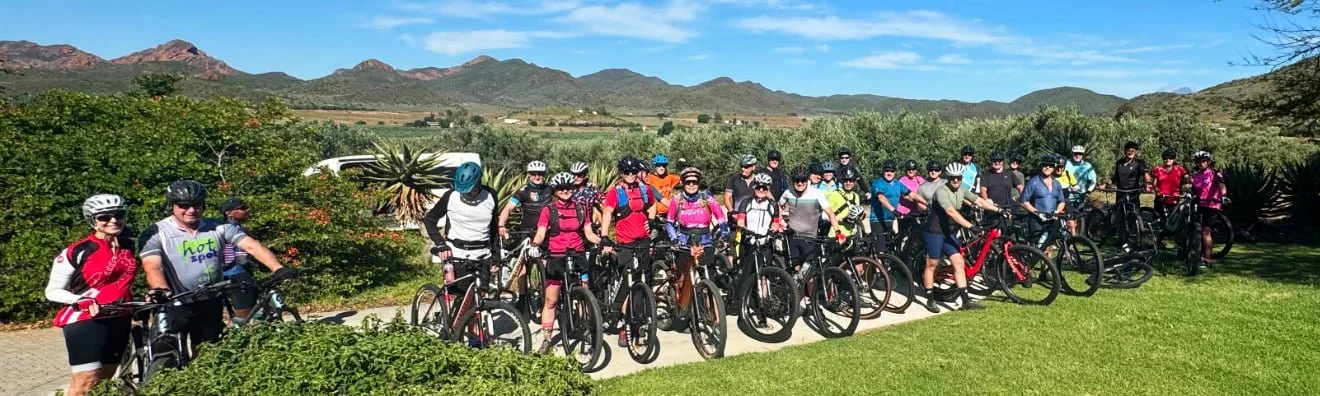 Group of people in cycling gear with helmets on a paved path in a scenic outdoor setting, with mountains and greenery in the background.