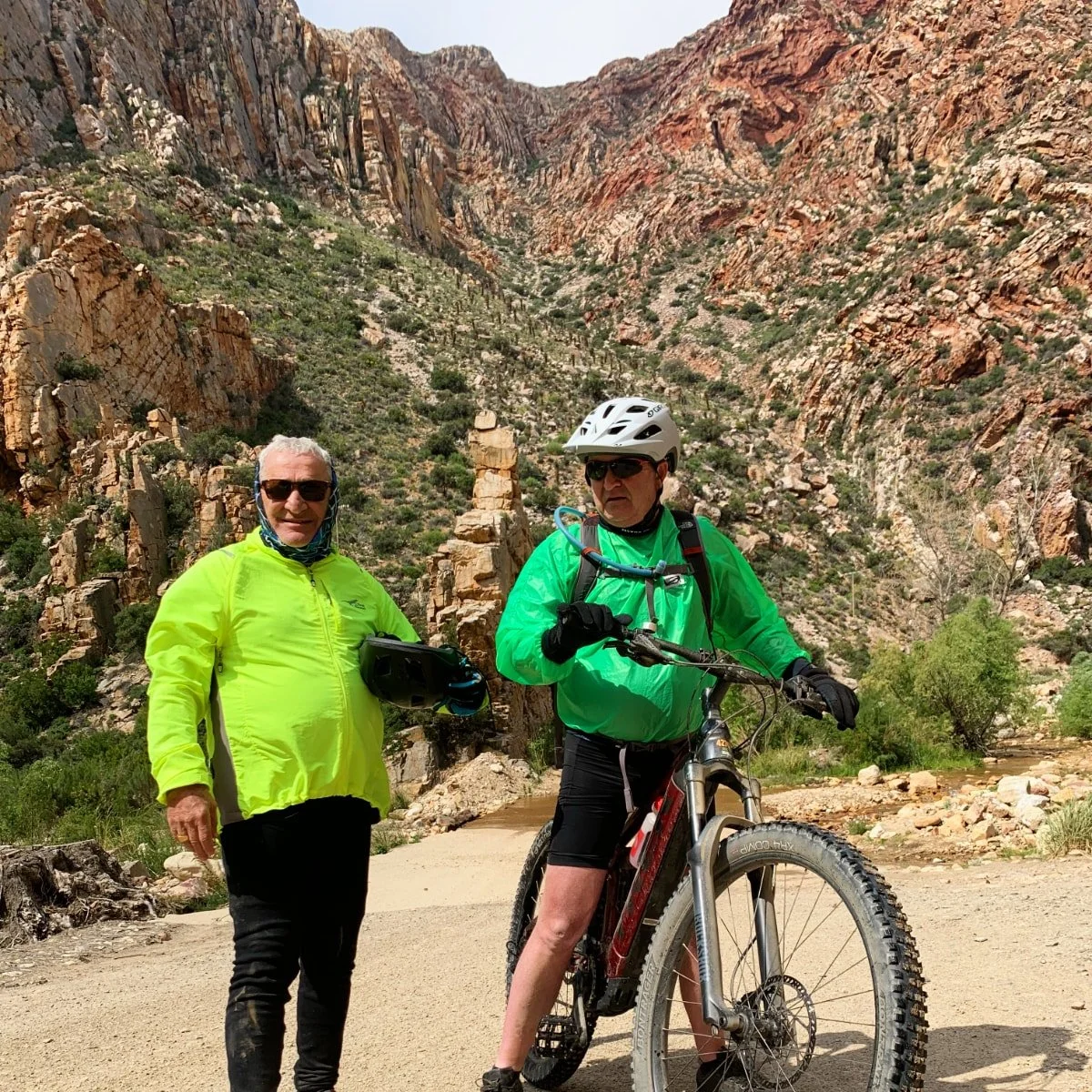 Two men in bright green jackets, one standing with a helmet in hand and the other on a mountain bike, in a desert canyon with rocky cliffs and sparse vegetation.