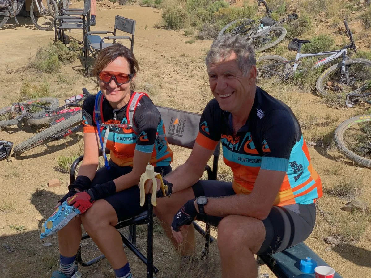 A woman and a man sitting on a bench in a desert area with several mountain bikes lying on the ground and leaning against each other in the background. They are dressed in cycling gear and smiling.