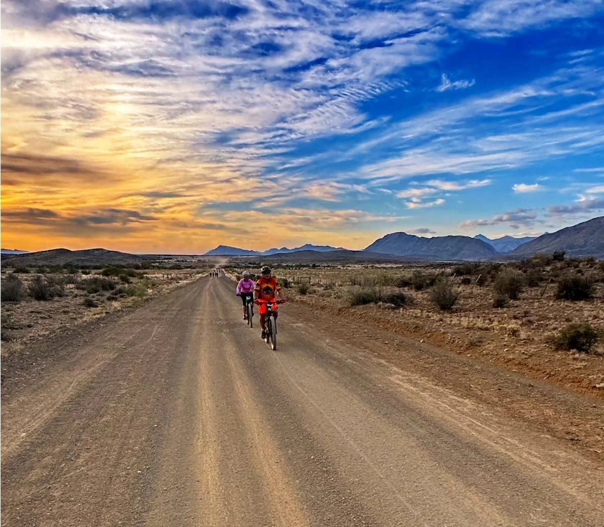 People biking on a dirt road in a desert landscape during sunset with mountains in the background and colorful sky.