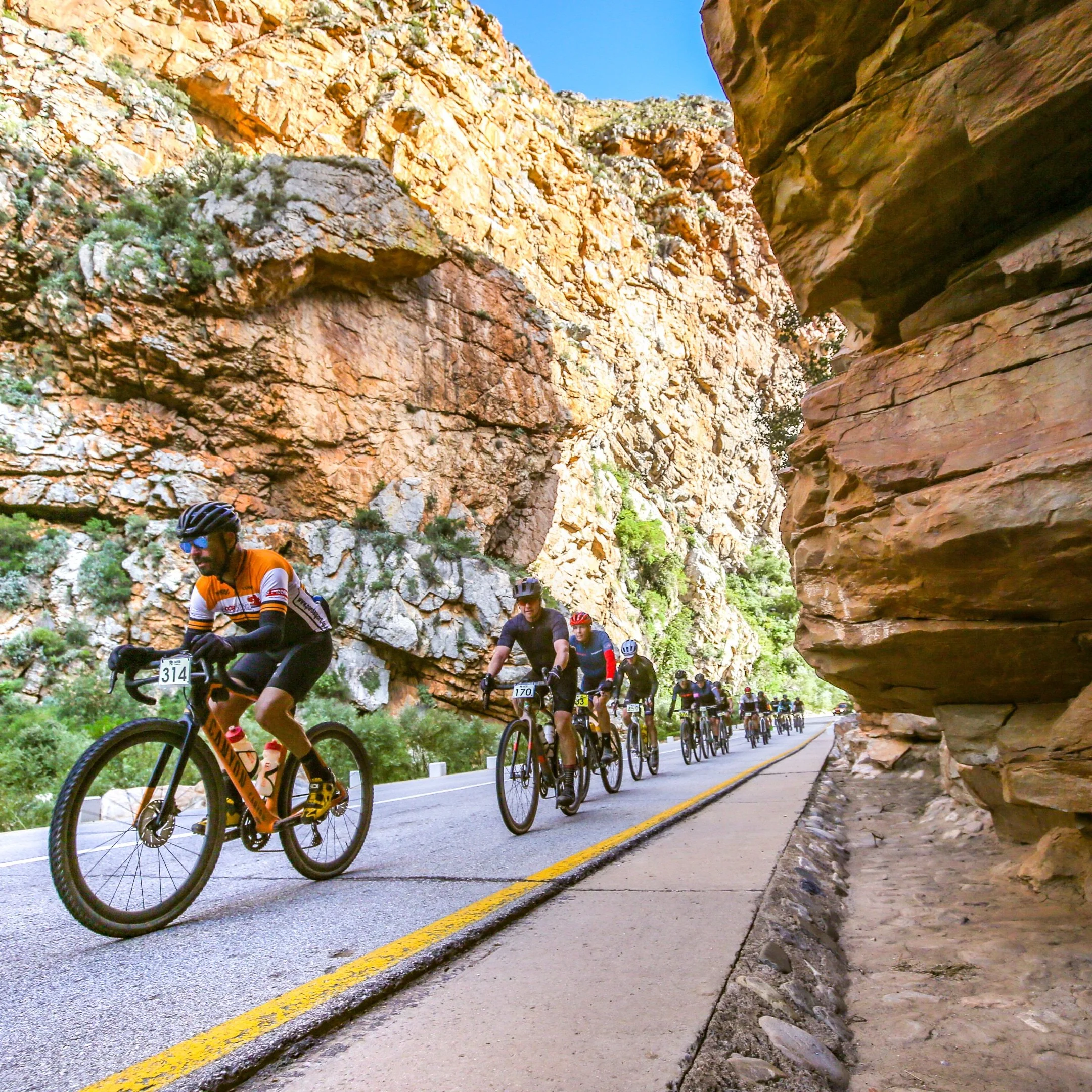A group of cyclists riding on a mountain road through a rugged, rocky canyon with tall, layered red and brown cliffs on the sides.