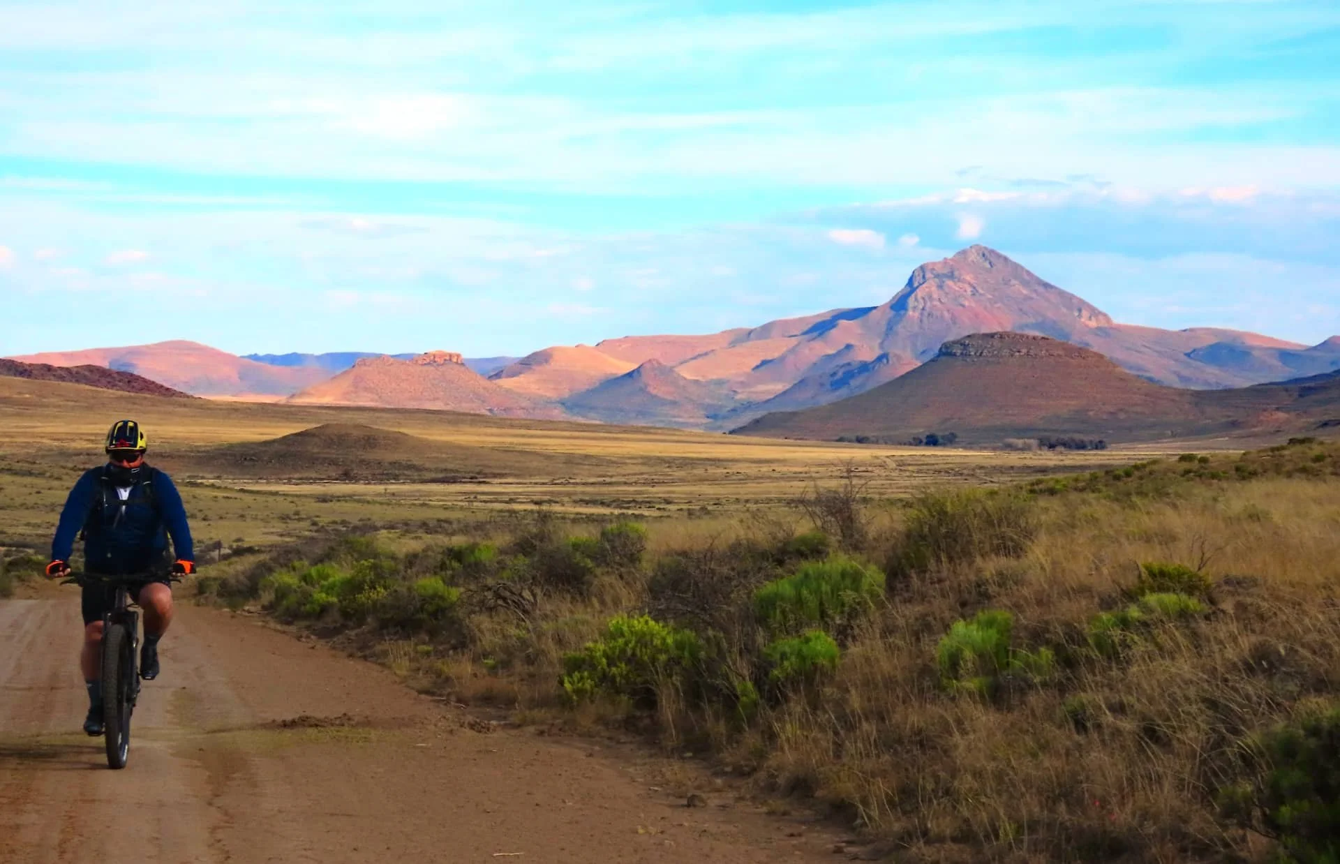 A person biking on a dirt trail through a landscape with mountains in the distance under a partly cloudy sky.