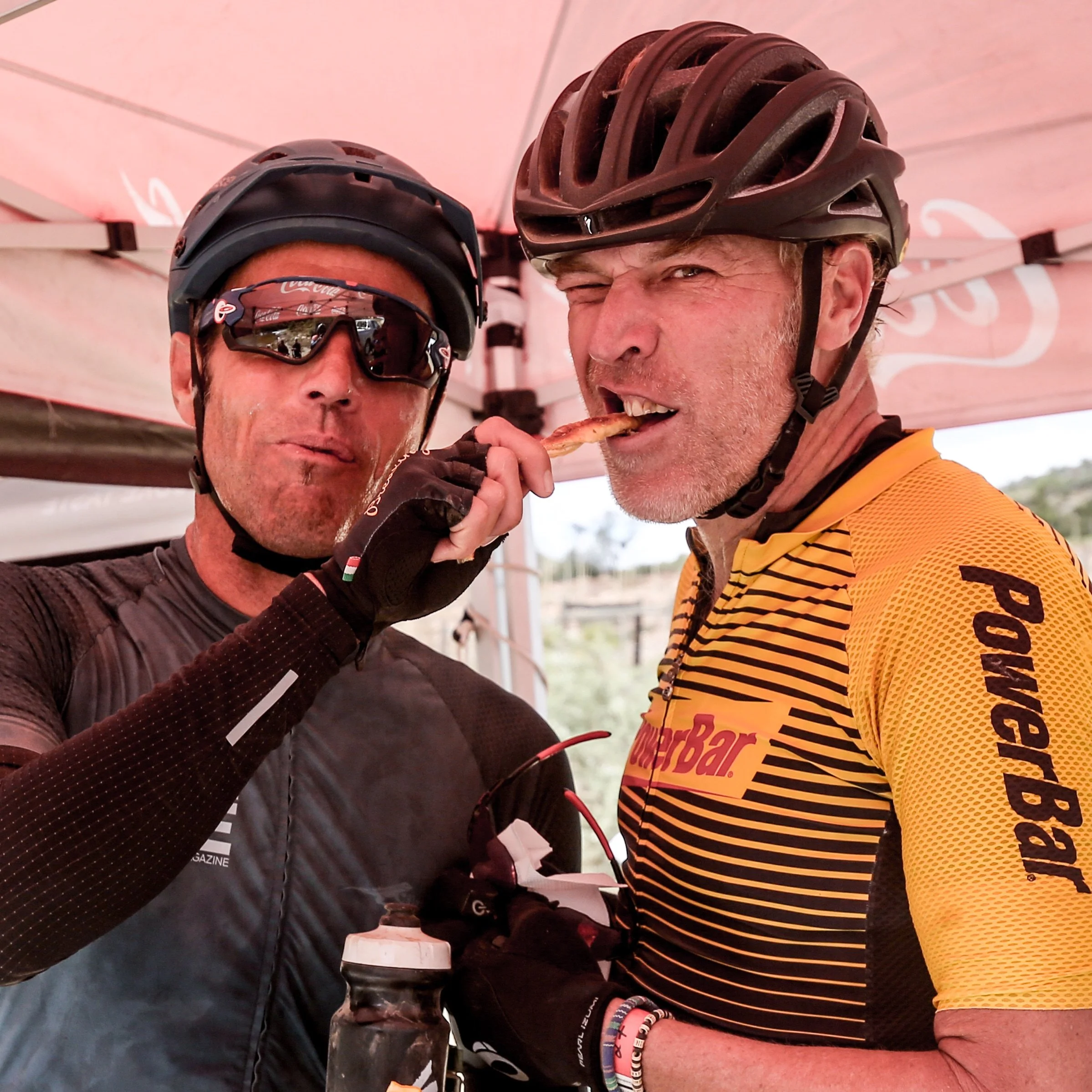 Two male cyclists, one wearing sunglasses and the other wearing a helmet, sharing a slice of pizza under a tent, dressed in cycling gear.