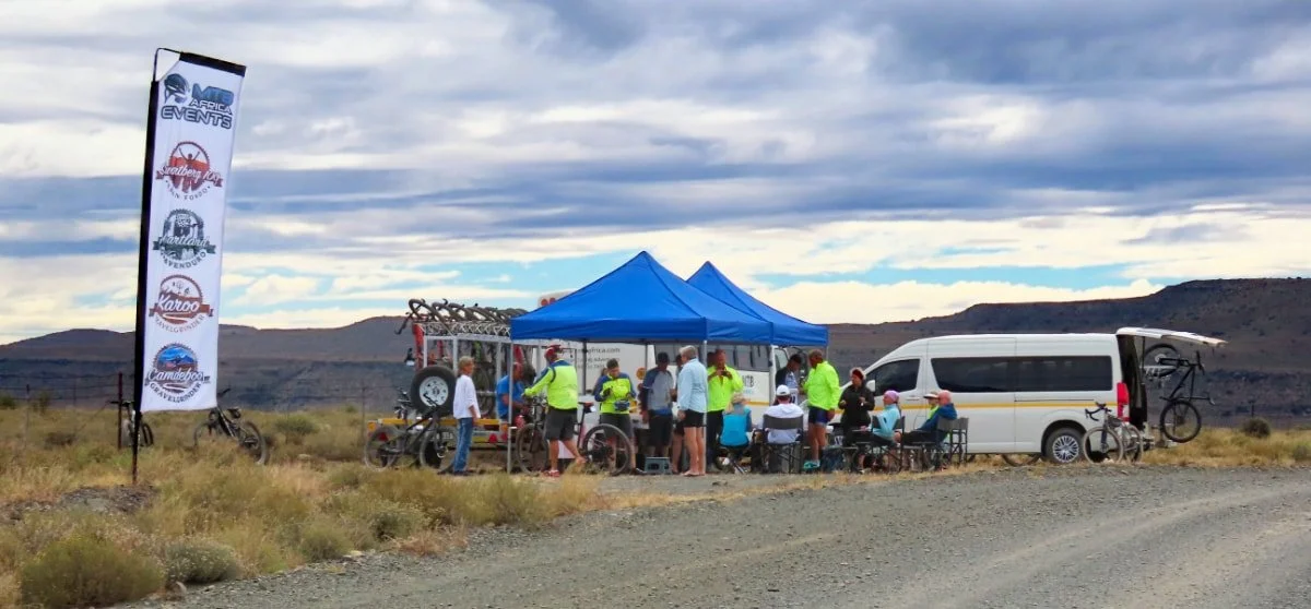 Group of people gathered under a blue canopy tent, with bikes and a white van parked nearby in a desert landscape.