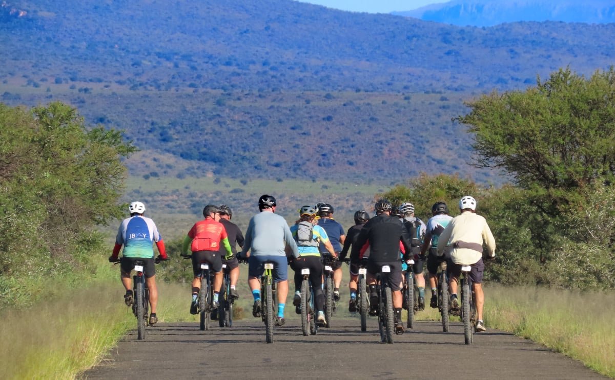 Group of people riding bicycles on a paved trail surrounded by greenery, mountains in the background.