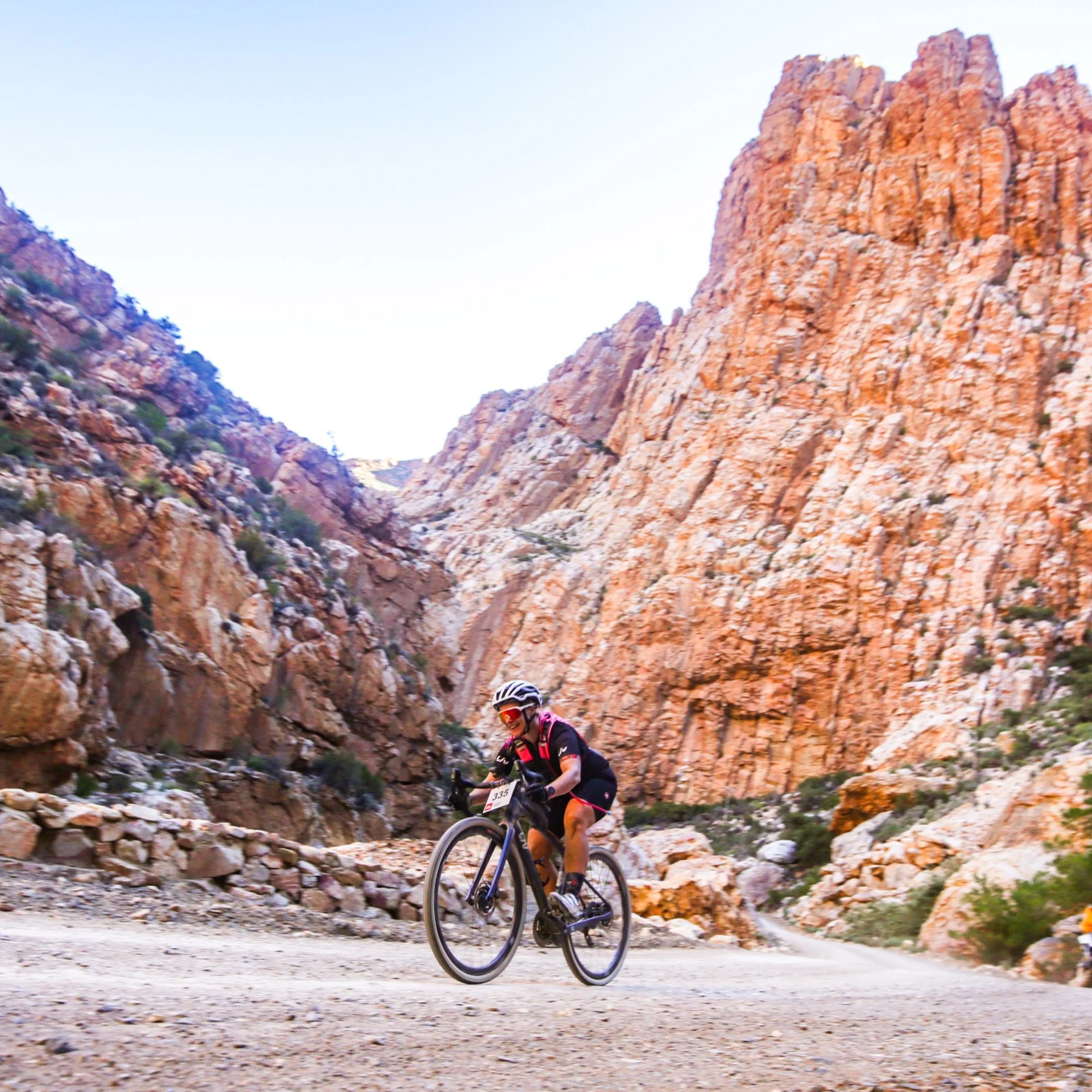 A cyclist riding on a dirt mountain trail surrounded by orange rocky cliffs, wearing a helmet and sports gear.