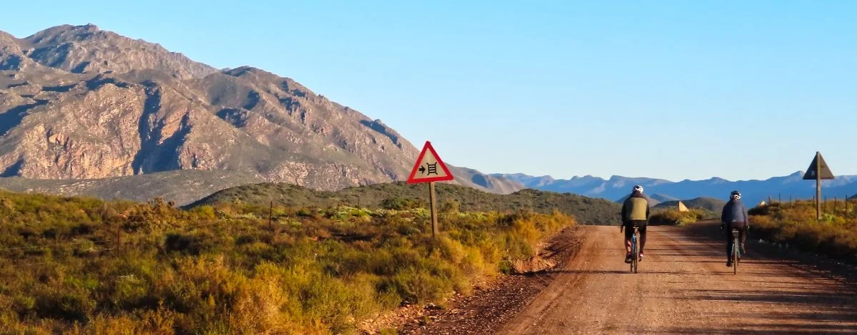 Two cyclists riding on a dirt road in a mountainous landscape with dry vegetation and a mountain range in the background.