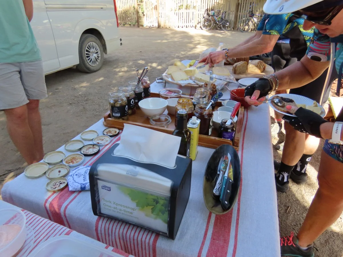 Outdoor picnic table set with bread, jams, and snacks, with people serving and preparing food under a shaded area near a fence and parked vehicles.