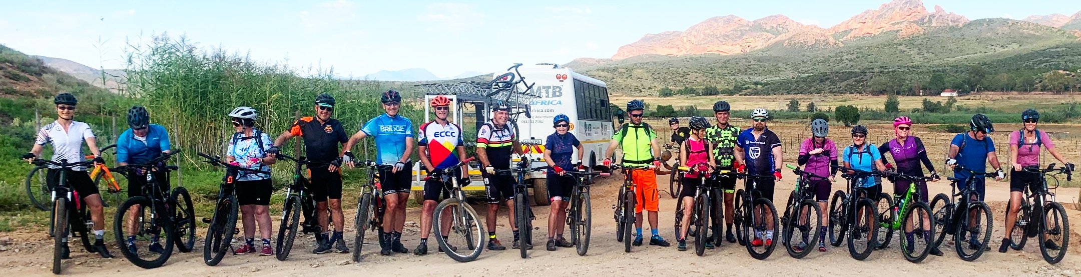 Group of cyclists standing with their bikes in an outdoor setting with mountains and a bus in the background.