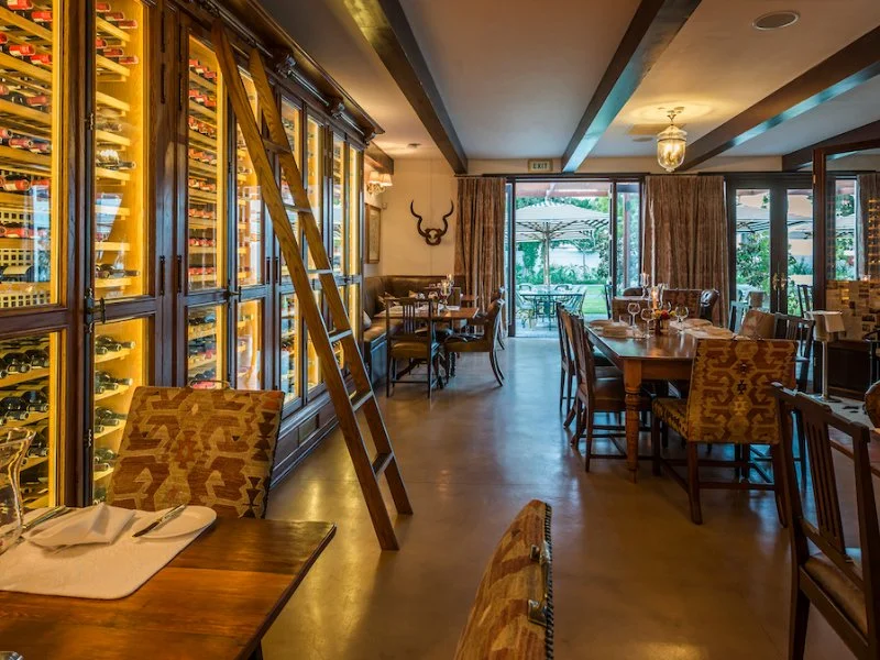 A cozy restaurant with wooden tables and chairs, large wine refrigerators along the left wall, a ladder leaning against the wine storage, and large windows with curtains revealing outdoor seating and greenery.