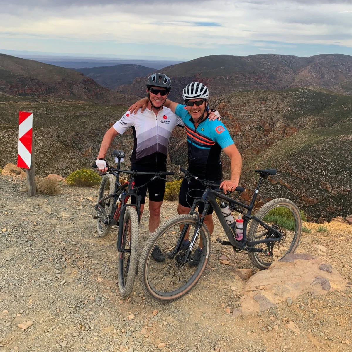 Two men wearing cycling gear and helmets smiling with their bicycles in a mountainous landscape with rocky terrain and cloudy sky.