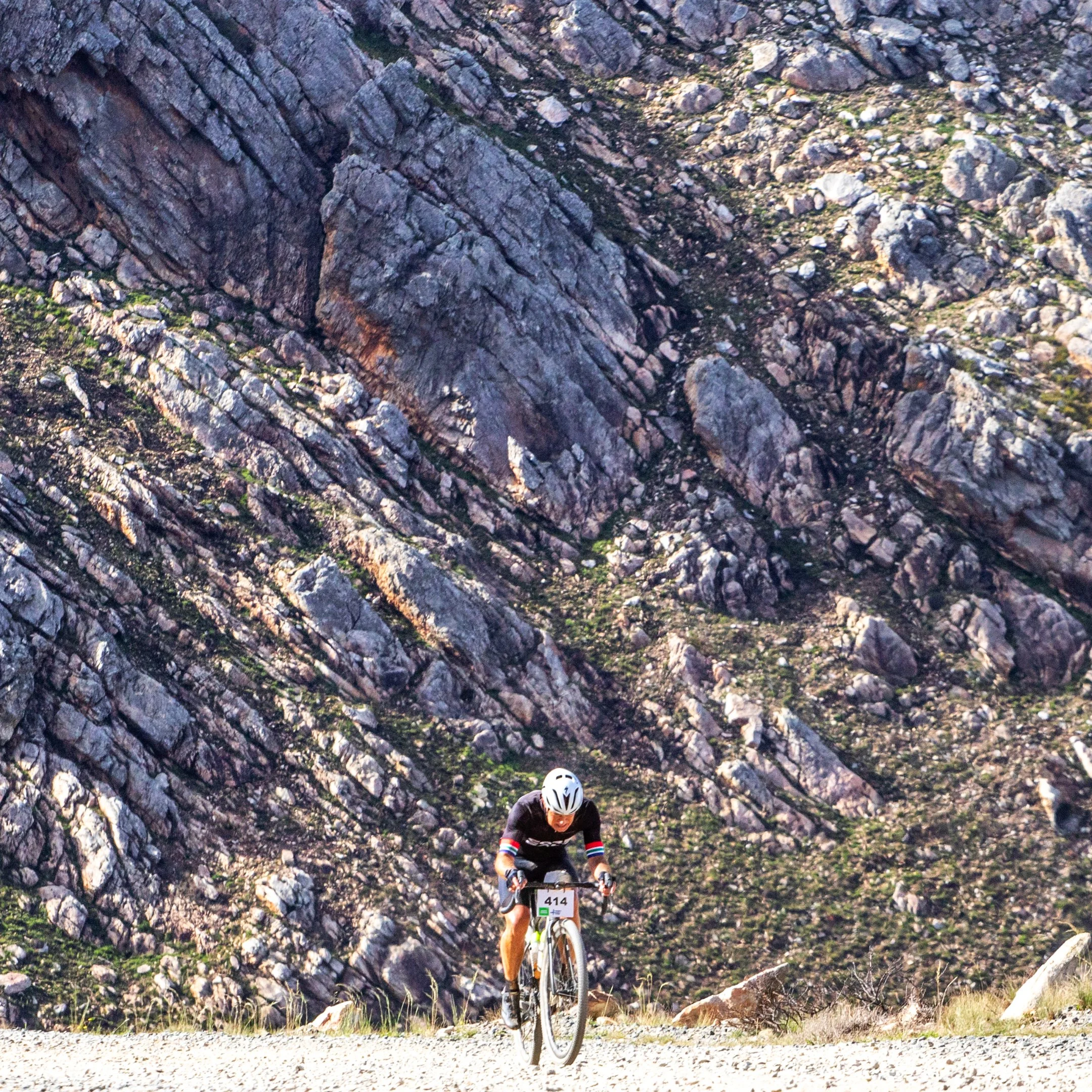A cyclist riding a mountain bike uphill on a gravel road against a rocky mountain backdrop.