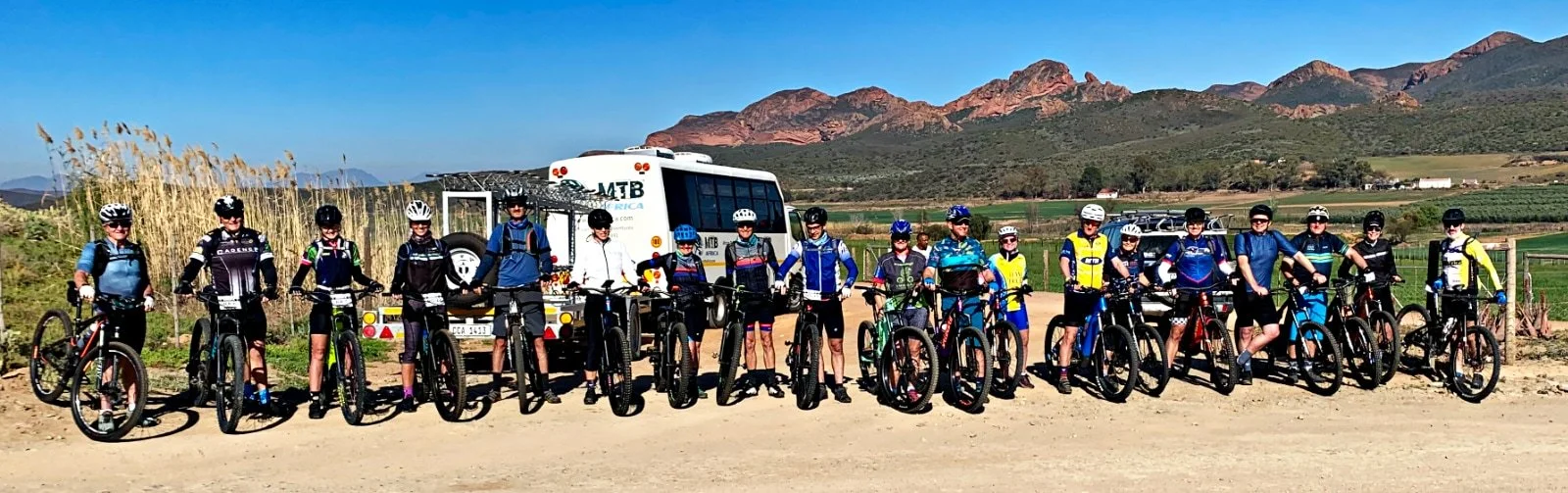 Group of people in cycling gear with helmets and sunglasses standing with bikes along a dirt road, mountain landscape in the background.