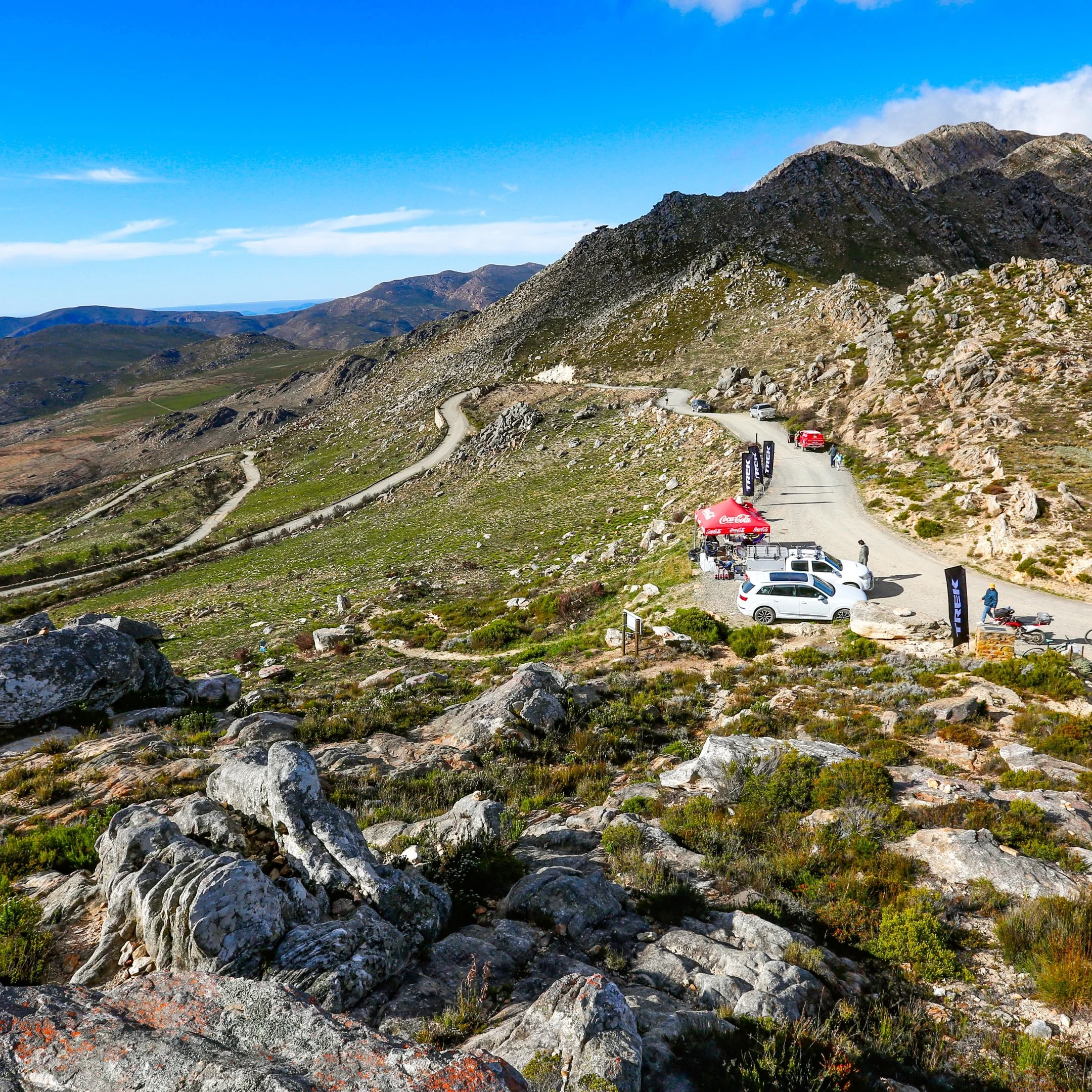 A mountain landscape with a winding dirt road, parked cars, a red tent, and several people.