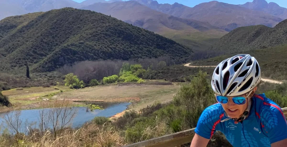 A cyclist wearing a white helmet, blue sunglasses, and a blue cycling jersey riding on a mountain road with hills, a river, and greenery in the background.