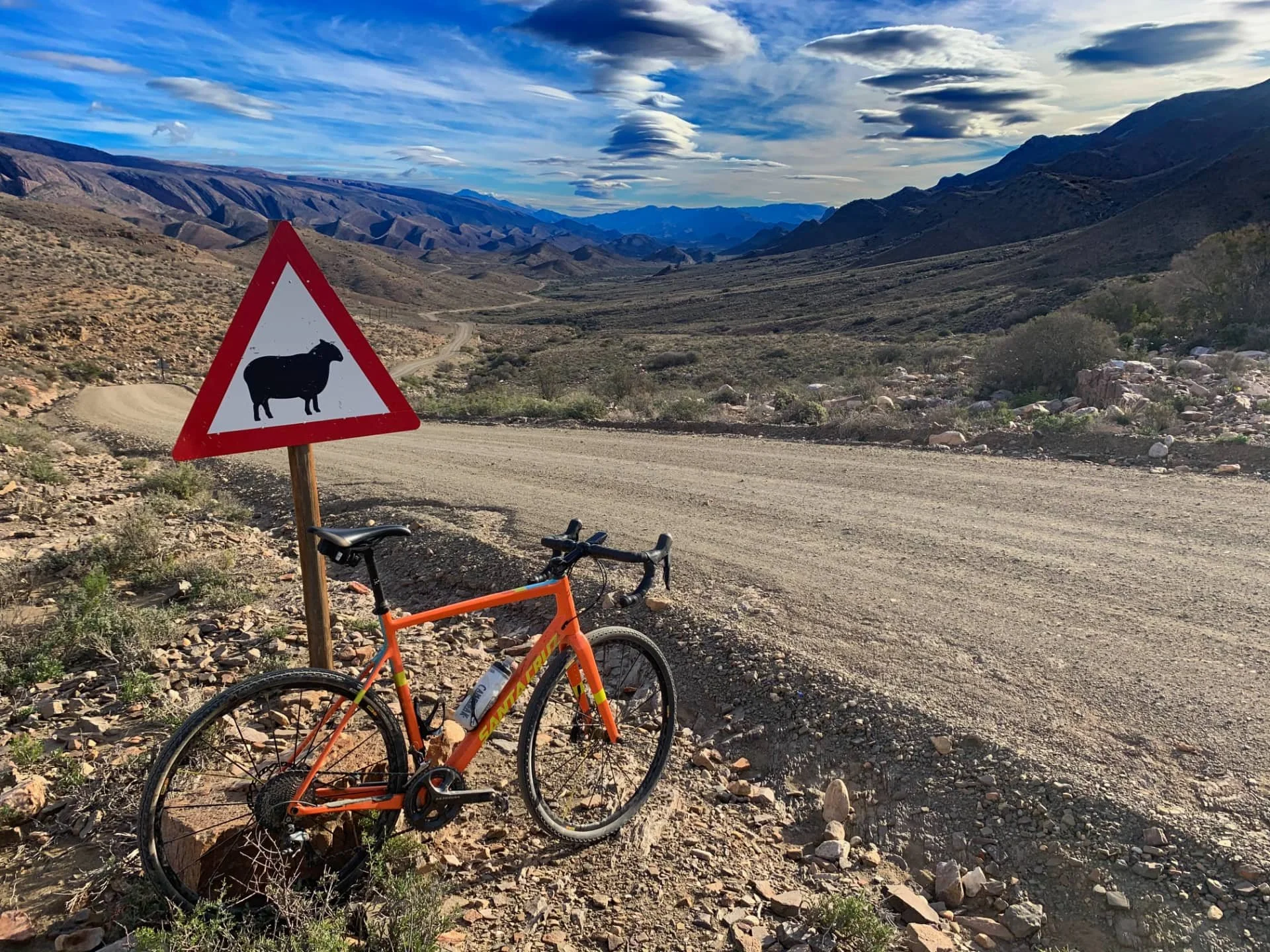 An orange bicycle parked on a dirt road next to a warning sign with a black sheep silhouette in a mountainous desert landscape.