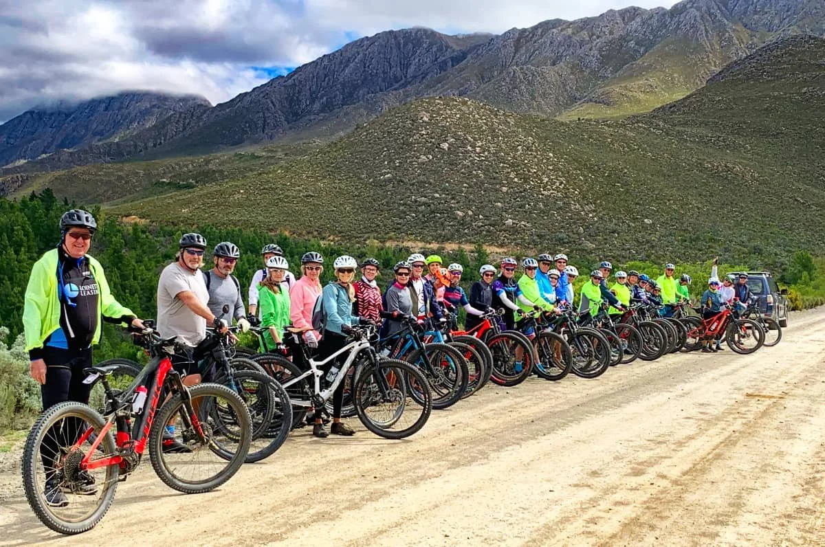 A group of people with mountain bikes lined up on a dirt trail, set against a backdrop of mountains and cloudy sky.