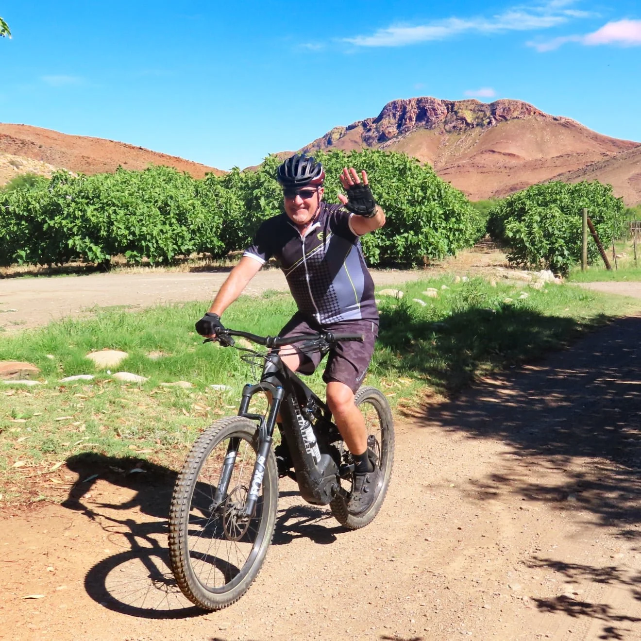 Man riding a mountain bike on a dirt trail, waving, with green trees and a mountain range in the background under a blue sky.