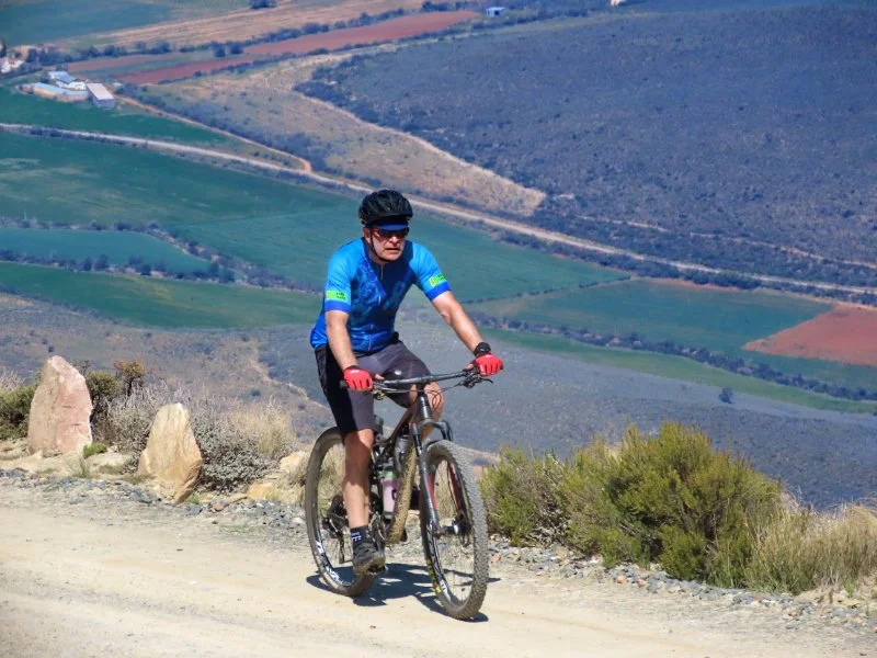 A man wearing sunglasses, a helmet, and a blue cycling jersey riding a mountain bike on a dirt trail in a hilly landscape with colorful fields and shrubs.